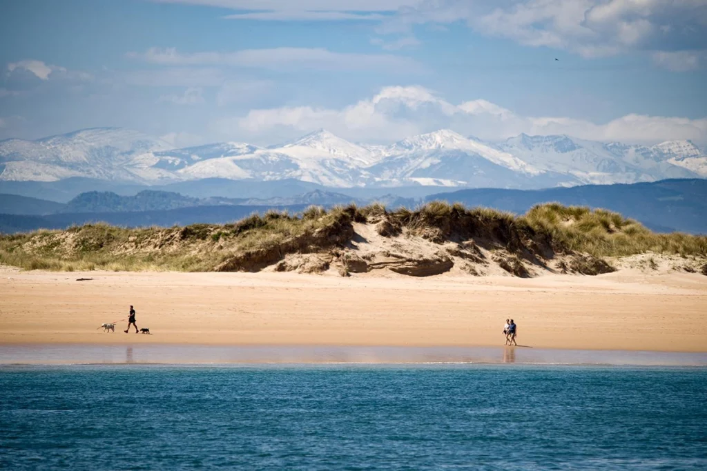 Personas pasean por playa con montañas nevadas de fondo